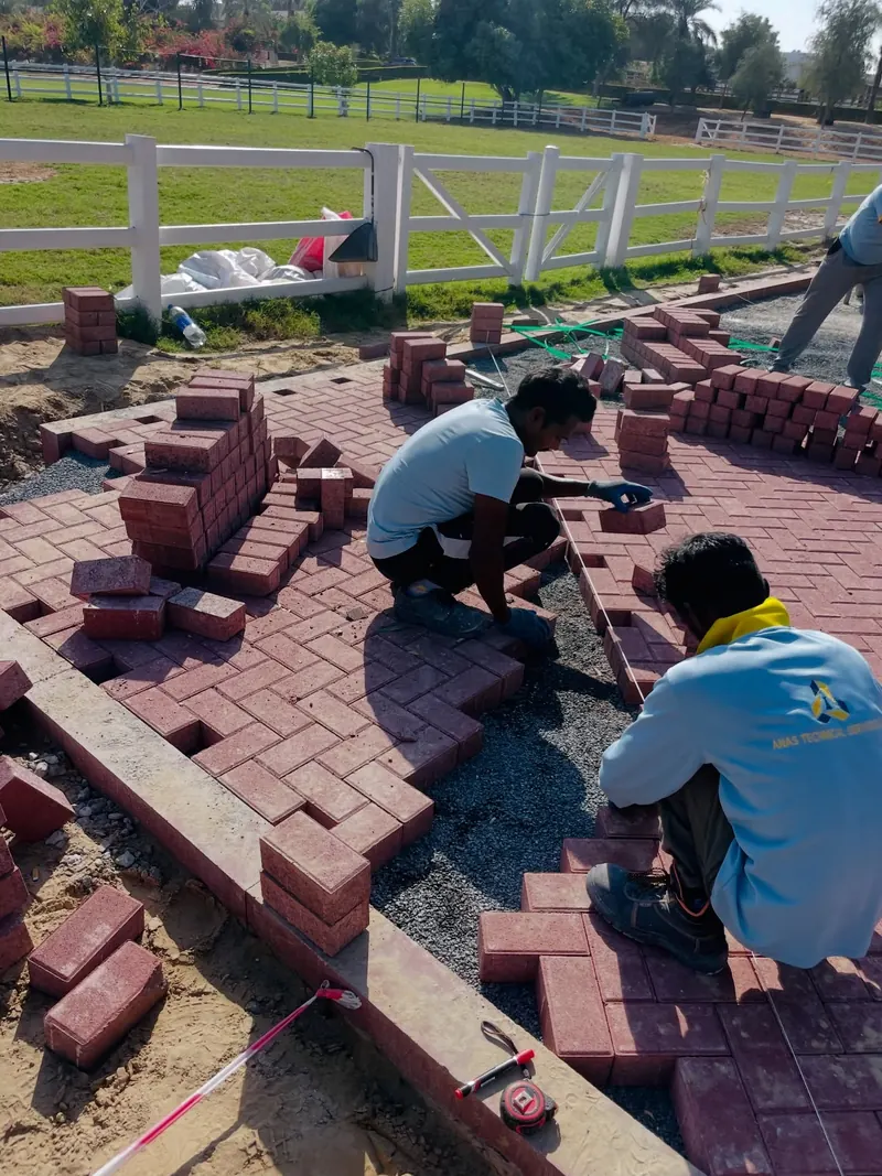 Two Anas Technical Services workers in company uniform laying red interlock pavers on a prepared sand bed, Dubai